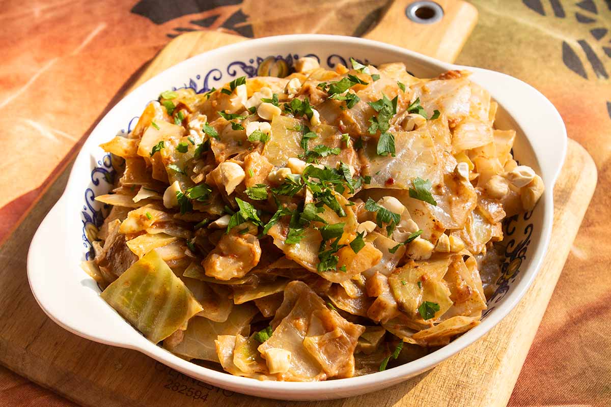 A white bowl filled with stir-fried cabbage and diced chicken, topped with chopped peanuts and fresh parsley. The dish is placed on a wooden board with a colorful tablecloth in the background.