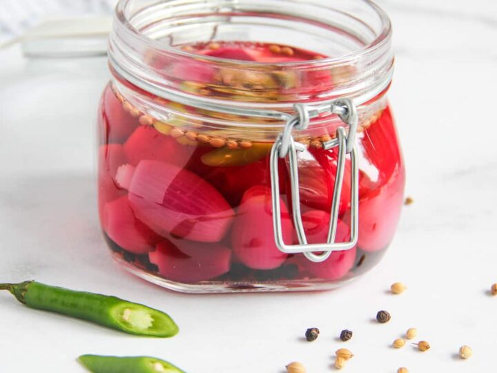A jar of pickled red onions with seeds in brine. The jar has a metal clasp and sits on a white surface. There are scattered coriander seeds, black peppercorns, and sliced green chili on the surface nearby.
