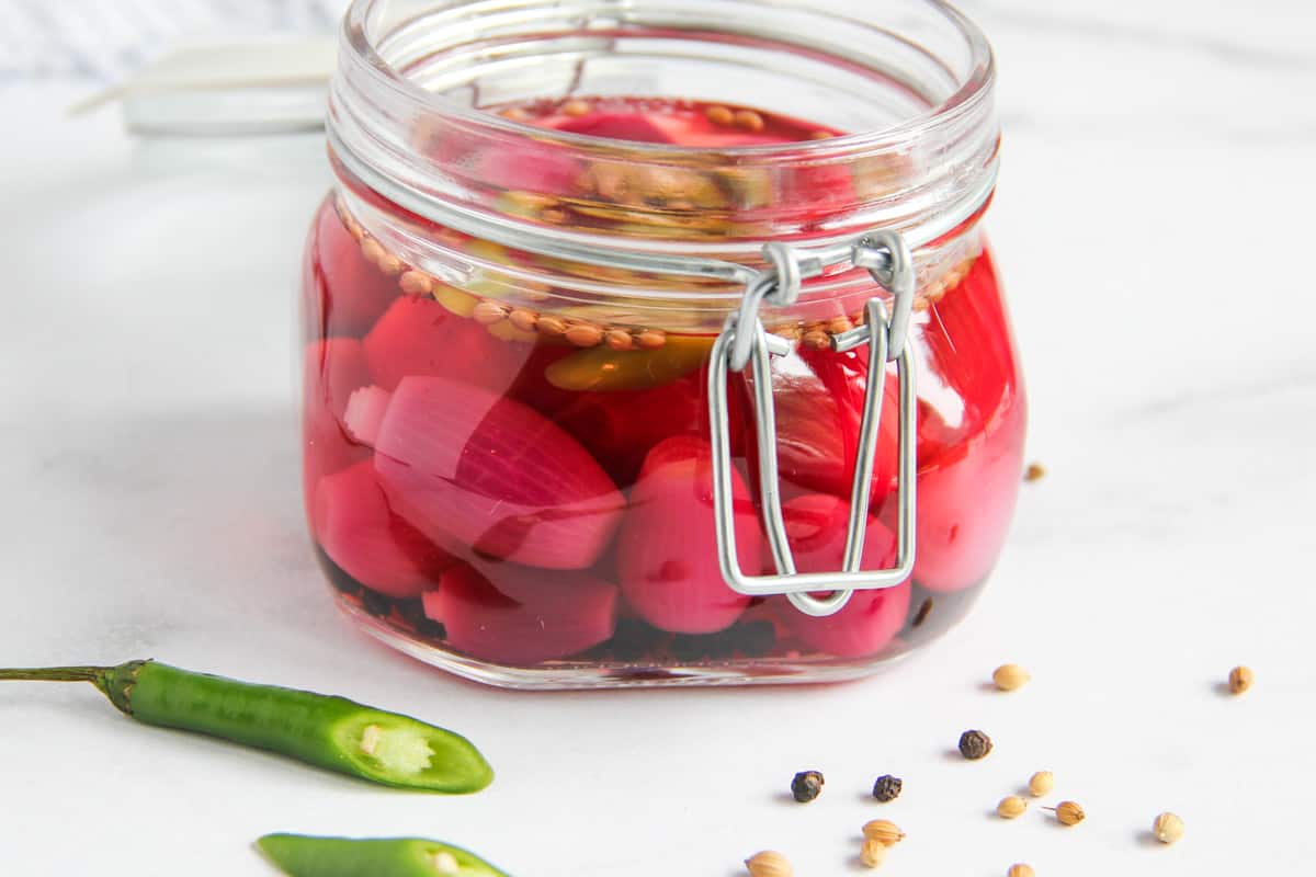 A jar of pickled red onions with seeds in brine. The jar has a metal clasp and sits on a white surface. There are scattered coriander seeds, black peppercorns, and sliced green chili on the surface nearby.