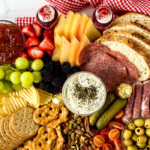 A picnic charcuterie board rests on a red and white checkered cloth, showcasing salami, bread, cheese, strawberries, grapes, blackberries, crackers, pretzels, nuts, cherry tomatoes, pickles, and cups of dip. Decorative text reads Easy Picnic Charcuterie Recipe.