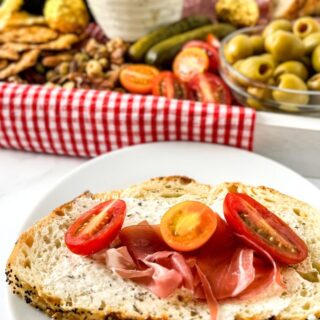 A slice of bread with cheese spread, prosciutto, and cherry tomato slices sits on a white plate. In the background, a vibrant picnic charcuterie board boasts olives, pickles, nuts, sliced meats, bread, and fruits atop a red checkered cloth.