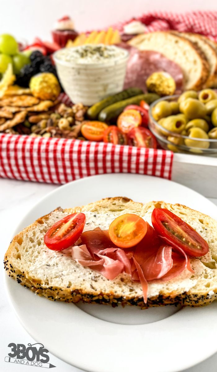 A slice of bread with cheese spread, prosciutto, and cherry tomato slices sits on a white plate. In the background, a vibrant picnic charcuterie board boasts olives, pickles, nuts, sliced meats, bread, and fruits atop a red checkered cloth.