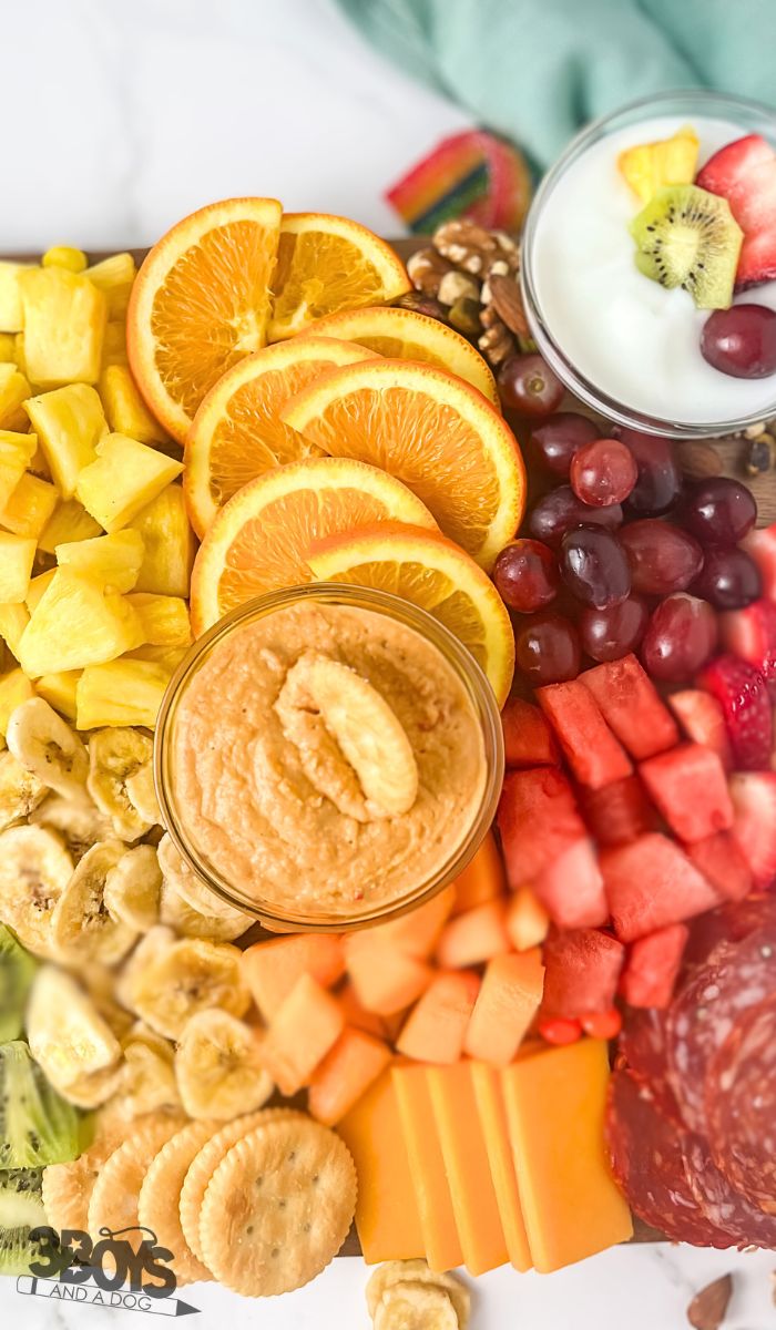 A vibrant rainbow charcuterie board showcases sliced oranges, pineapple, bananas, watermelon, and strawberries. It comes complete with grapes, kiwi, salami, cheddar cheese, crackers, a bowl of hummus, and a cup of yogurt topped with fruit. A feast for the eyes and palate!.