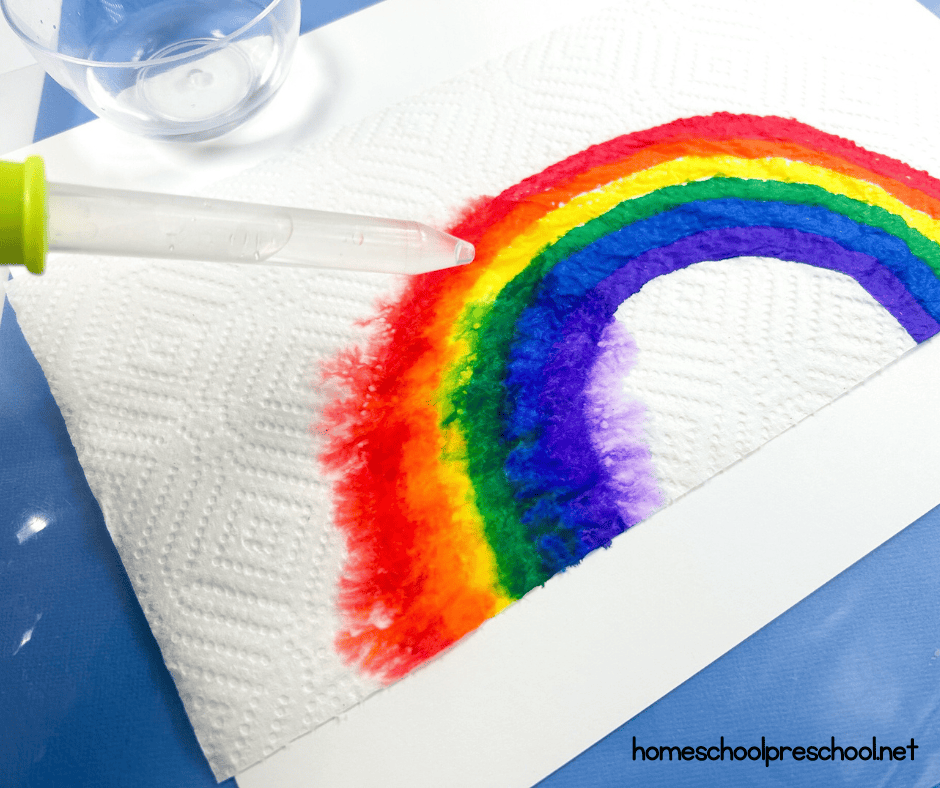 A vibrant rainbow, painted spread on a sheet of paper towel, with a dropper nearby. The rainbows colors blend slightly, and a clear plastic cup sits in the top left corner. The scene suggests a fun, creative art activity.
