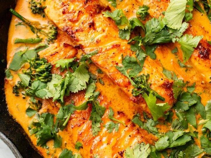 A skillet with creamy orange curry sauce containing pieces of cooked salmon. The dish is garnished with fresh cilantro and lime wedges. Broccoli florets are also visible in the sauce.