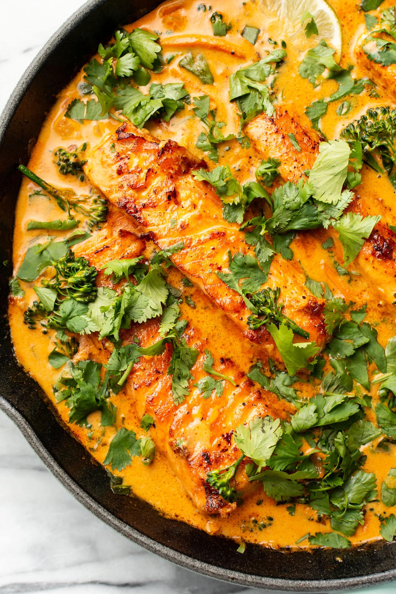 A skillet with creamy orange curry sauce containing pieces of cooked salmon. The dish is garnished with fresh cilantro and lime wedges. Broccoli florets are also visible in the sauce.