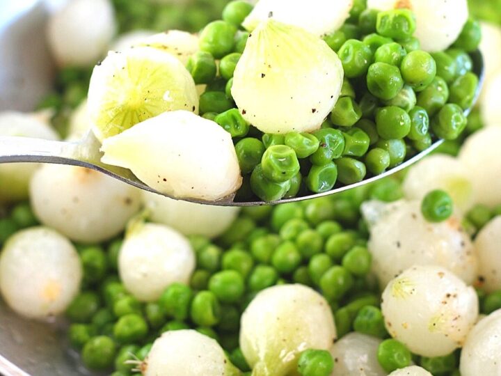 A spoon holding a serving of cooked peas and pearl onions over a bowl filled with more peas and onions. The vegetables are seasoned, with some black pepper visible on the surface.