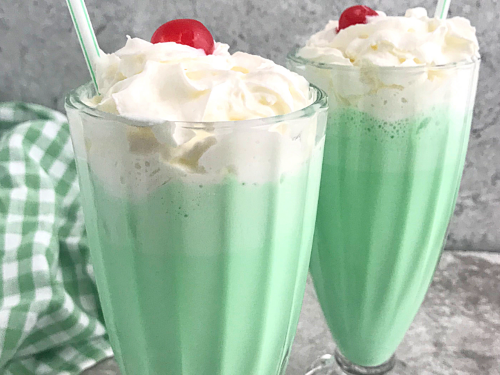 Two tall glasses of green milkshakes topped with whipped cream and a cherry. Each has a plastic straw. A checkered green cloth is in the background on a gray marble surface.