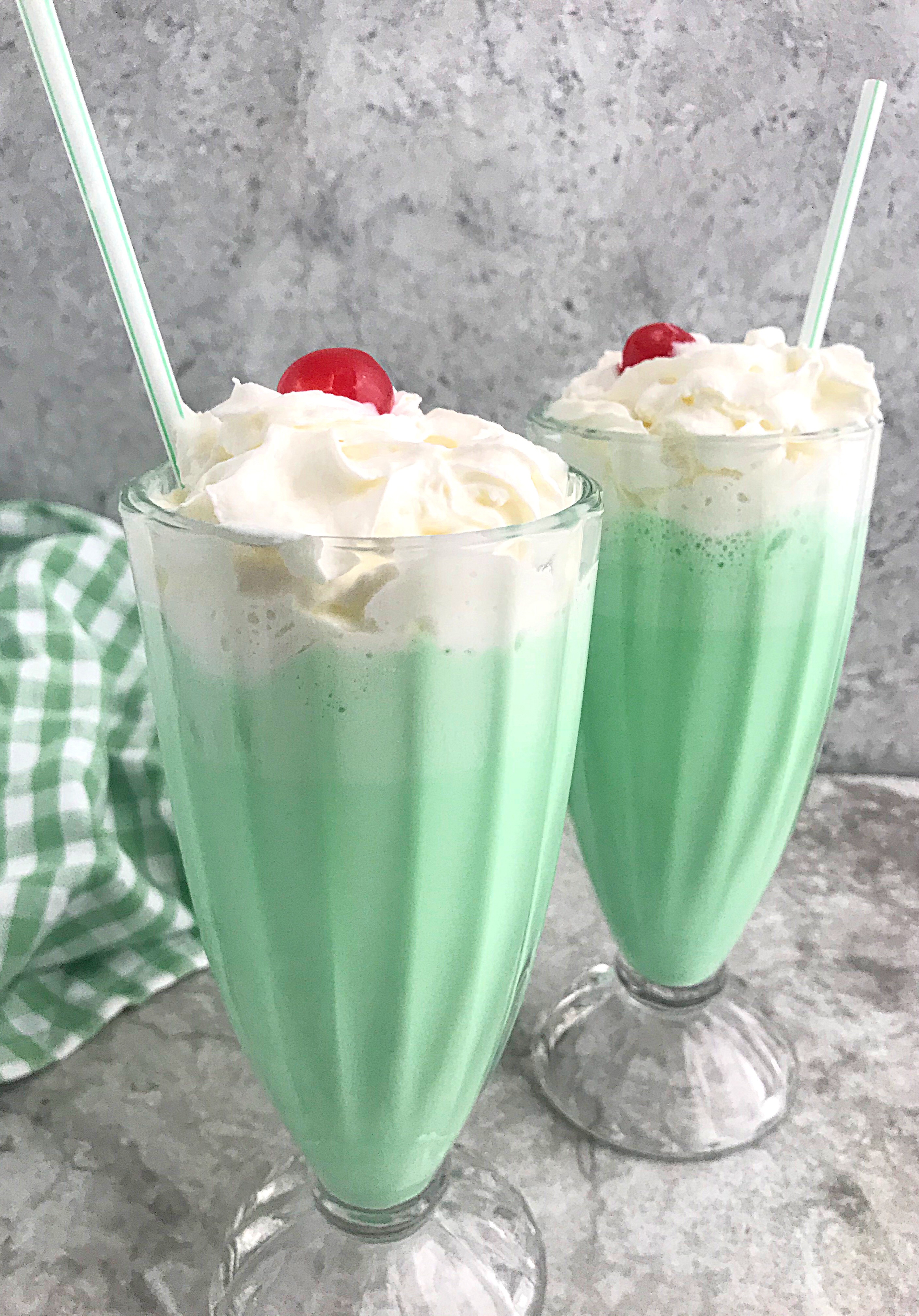 Two tall glasses of green milkshakes topped with whipped cream and a cherry. Each has a plastic straw. A checkered green cloth is in the background on a gray marble surface.
