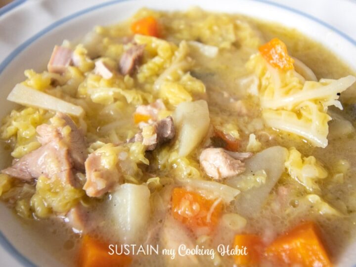 Close-up of a bowl of chicken soup containing chunks of chicken, carrots, potatoes, cabbage, and a clear broth. The soup sits in a white bowl with a blue rim. A spoon is partially visible in the soup.