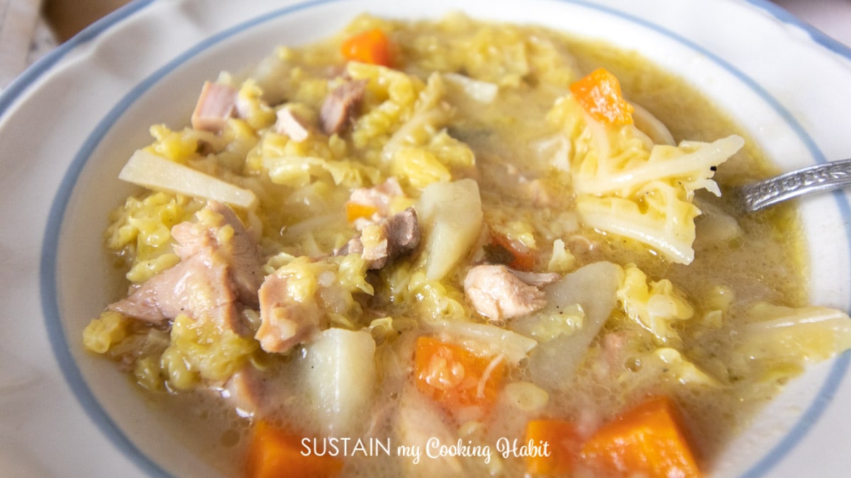Close-up of a bowl of chicken soup containing chunks of chicken, carrots, potatoes, cabbage, and a clear broth. The soup sits in a white bowl with a blue rim. A spoon is partially visible in the soup.