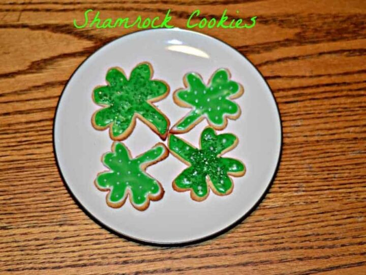 Four shamrock-shaped cookies with green icing are arranged on a white plate. The background is a wooden table, and the text Shamrock Cookies is written above in green.