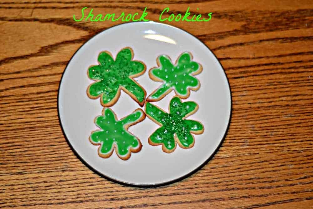 Four shamrock-shaped cookies with green icing are arranged on a white plate. The background is a wooden table, and the text Shamrock Cookies is written above in green.