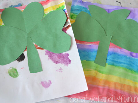 Two pieces of paper display colorful backgrounds; one with pastel rainbow stripes and the other with multicolored paint splatters. Each paper has a green paper shamrock glued on top.
