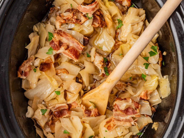 A slow cooker filled with cooked cabbage, pieces of bacon, and garnished with chopped parsley. A wooden spoon is resting inside the cooker. Fresh parsley leaves are visible in the top left corner.