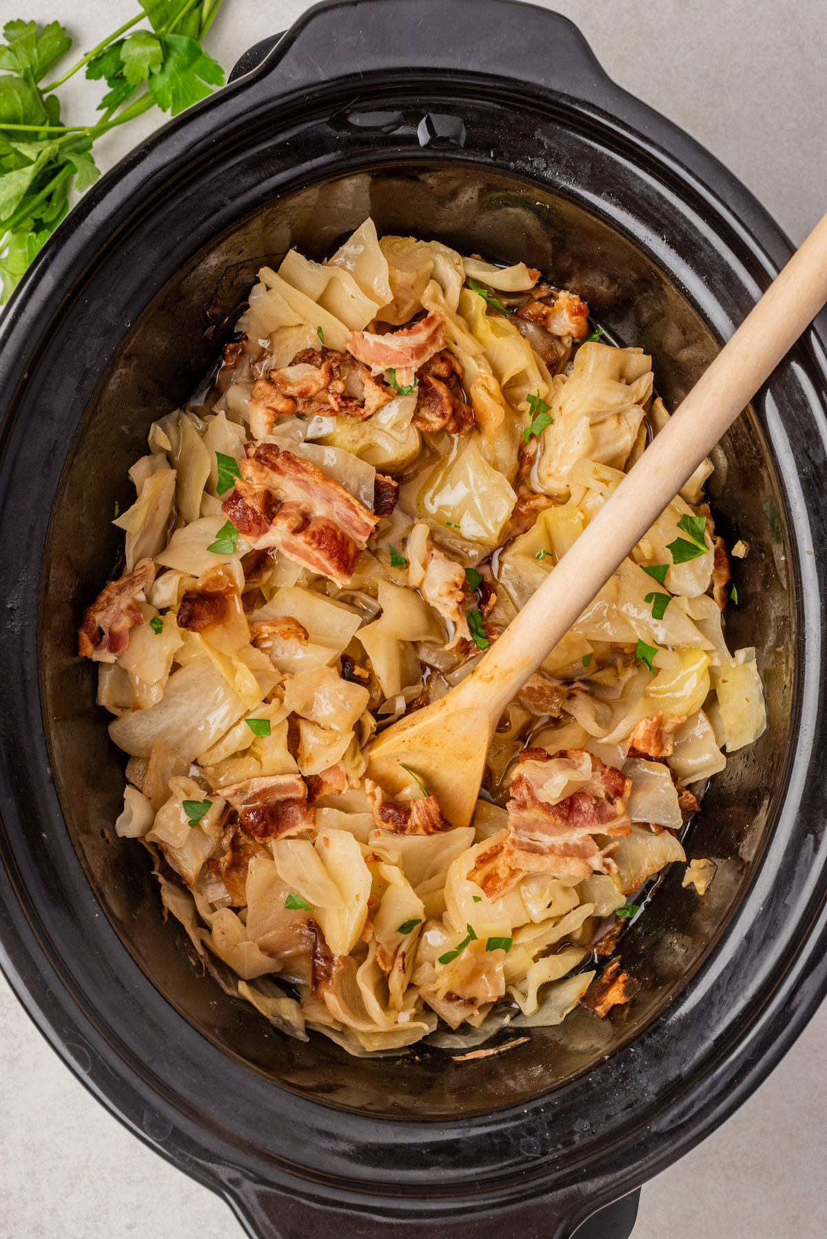 A slow cooker filled with cooked cabbage, pieces of bacon, and garnished with chopped parsley. A wooden spoon is resting inside the cooker. Fresh parsley leaves are visible in the top left corner.