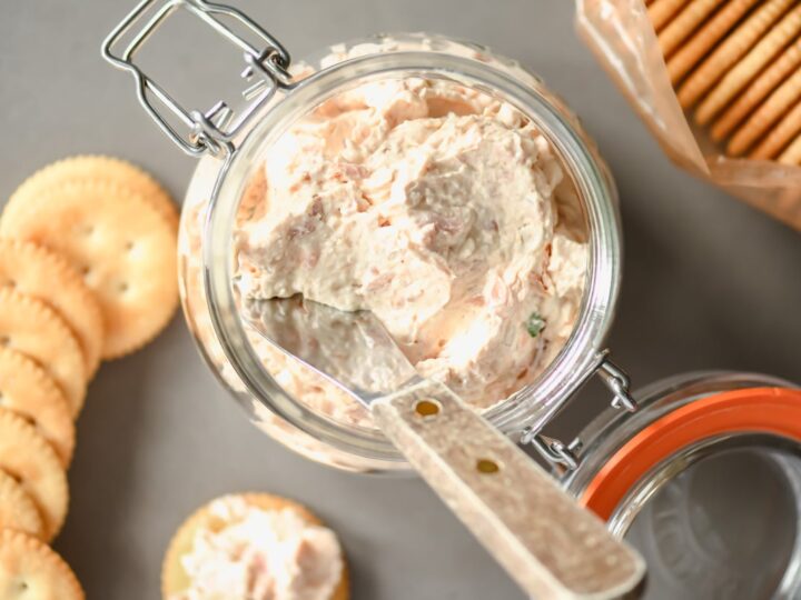 Glass jar filled with a creamy spread on a table, next to a spreading knife. Round crackers are placed in a scattered arrangement beside the jar, with some more crackers visible inside an open plastic sleeve.