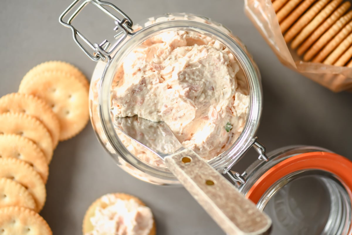 Glass jar filled with a creamy spread on a table, next to a spreading knife. Round crackers are placed in a scattered arrangement beside the jar, with some more crackers visible inside an open plastic sleeve.