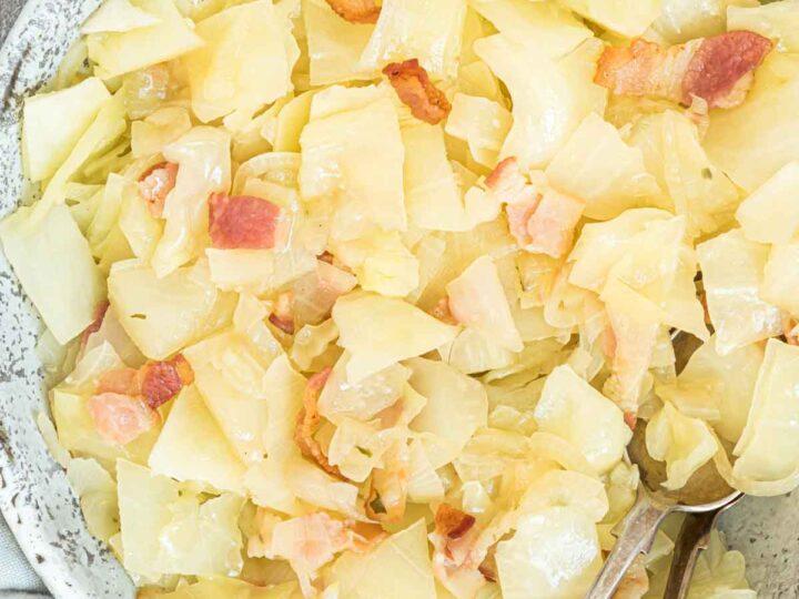 A speckled bowl filled with cooked cabbage and chunks of bacon. Two spoons rest on the side of the bowl, which is placed on a textured gray surface. The cabbage appears to be sautéed, showcasing a glossy texture.
