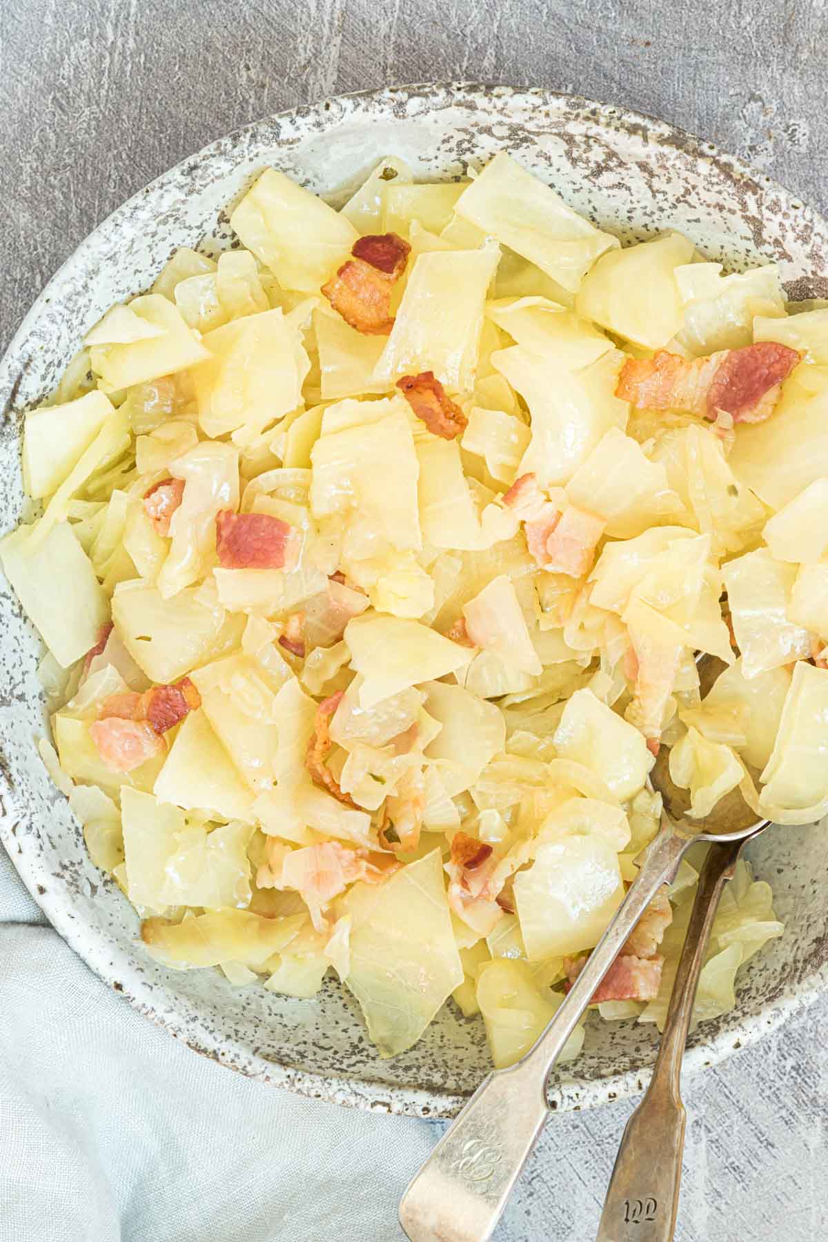 A speckled bowl filled with cooked cabbage and chunks of bacon. Two spoons rest on the side of the bowl, which is placed on a textured gray surface. The cabbage appears to be sautéed, showcasing a glossy texture.