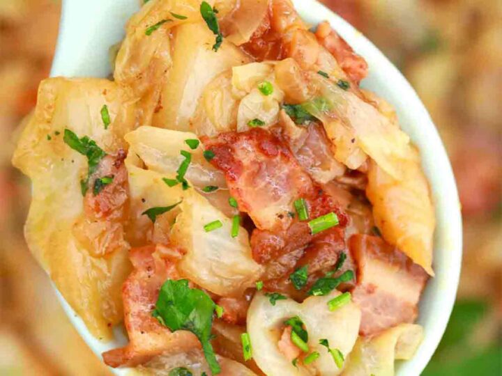 A close-up of a white spoon filled with a dish made of chopped cabbage, crispy bacon, and garnished with fresh green herbs. The background is blurred, showing more of the same dish.
