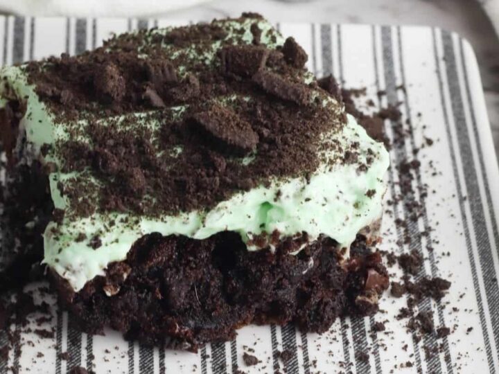 A piece of chocolate cake topped with mint green frosting and crumbled chocolate cookies is placed on a striped white and black plate.