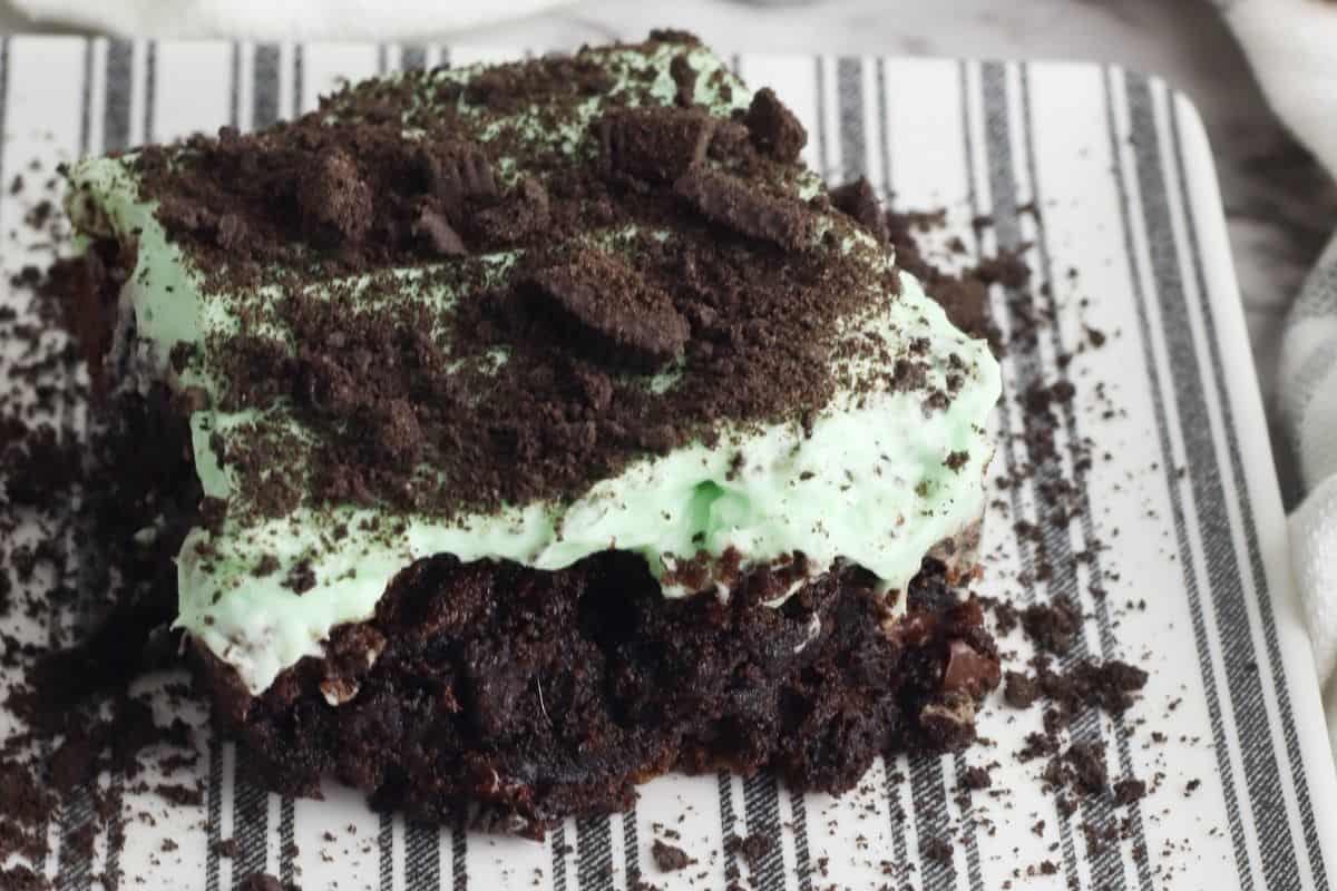 A piece of chocolate cake topped with mint green frosting and crumbled chocolate cookies is placed on a striped white and black plate.