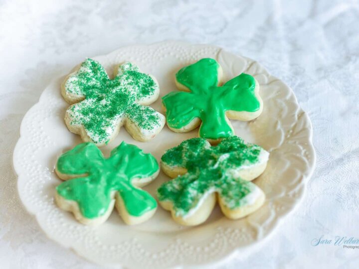 A white plate with four shamrock-shaped cookies on a lace-patterned tablecloth. Two cookies are decorated with green glittery sprinkles, and two are frosted with green icing.