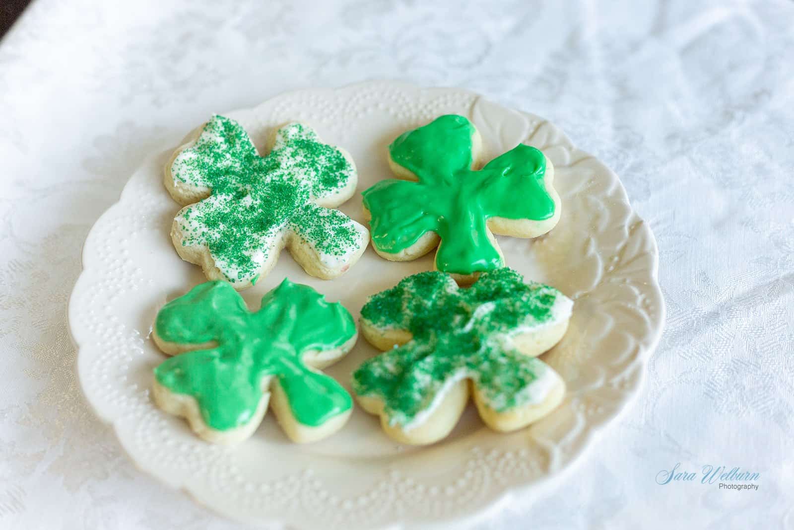 A white plate with four shamrock-shaped cookies on a lace-patterned tablecloth. Two cookies are decorated with green glittery sprinkles, and two are frosted with green icing.