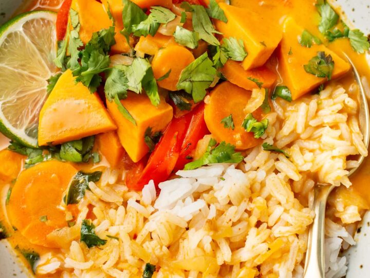 A bowl of creamy curry with carrots, red peppers, and chunks of sweet potato, garnished with cilantro and lime slices. Served with rice on the side in a speckled ceramic bowl, with a spoon resting on the side.