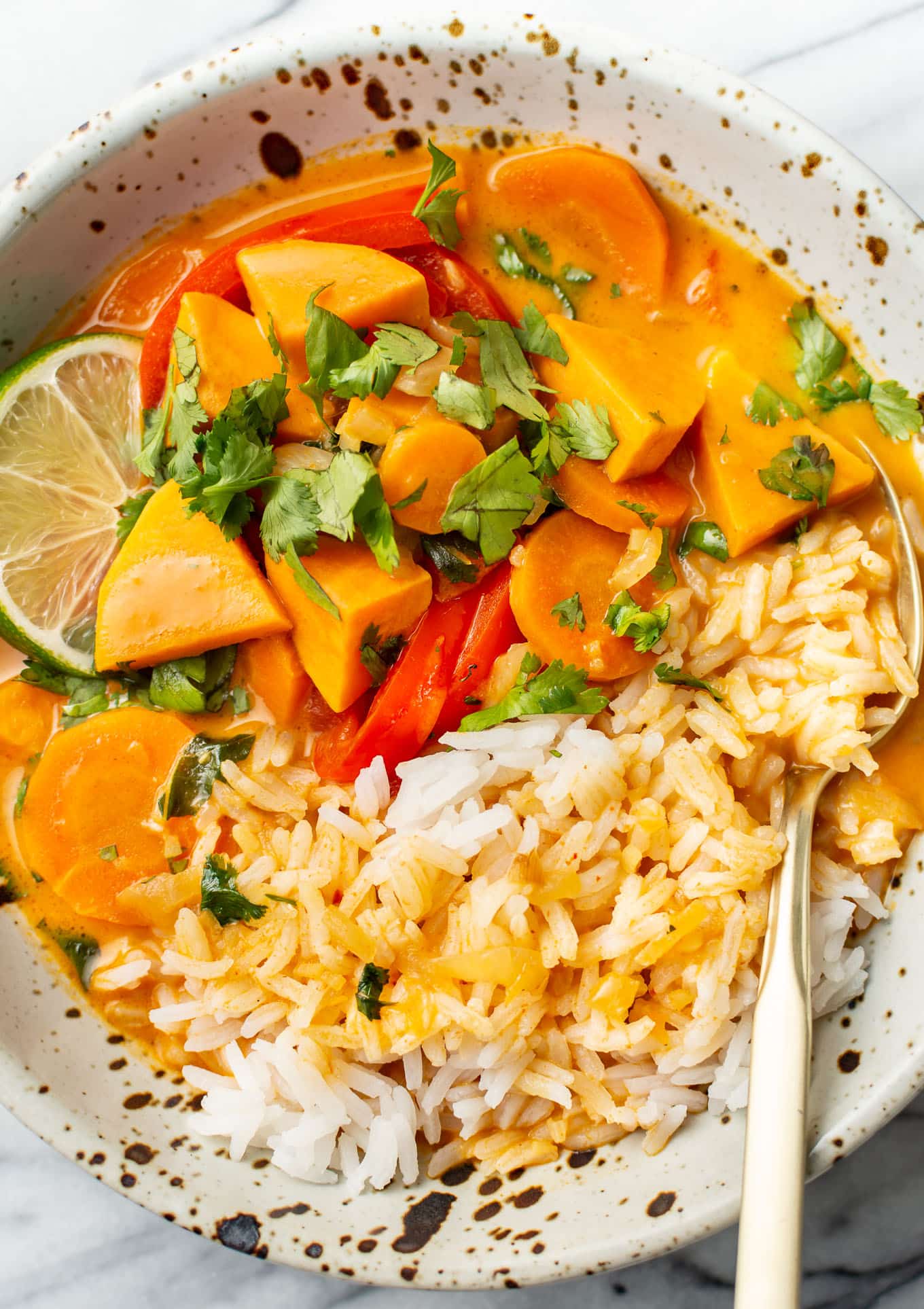 A bowl of creamy curry with carrots, red peppers, and chunks of sweet potato, garnished with cilantro and lime slices. Served with rice on the side in a speckled ceramic bowl, with a spoon resting on the side.