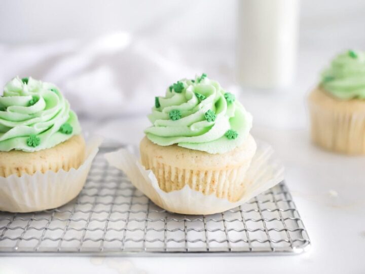 Two vanilla cupcakes with light green frosting and green sprinkles sit on a wire rack. A blurred cupcake and a bottle of milk are in the background on a white surface.