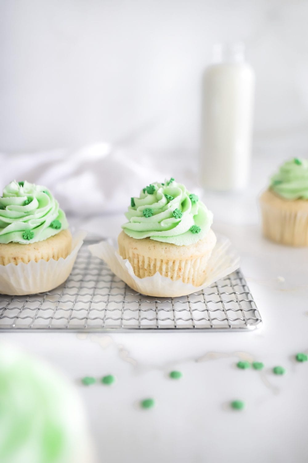 Two vanilla cupcakes with light green frosting and green sprinkles sit on a wire rack. A blurred cupcake and a bottle of milk are in the background on a white surface.