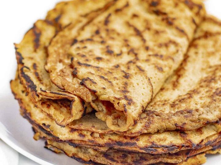A stack of golden-brown keto tortillas on a white plate, showing a slightly crispy texture. The top tortilla is slightly folded, highlighting the soft interior. The background is white, emphasizing the tortillas.