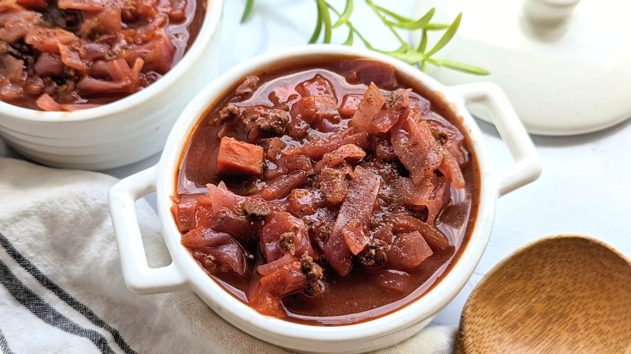 A white bowl filled with a hearty stew featuring chunks of beef and cabbage in a rich tomato-based broth. Another bowl is partially visible in the background, along with a sprig of fresh herbs and a wooden spoon nearby.