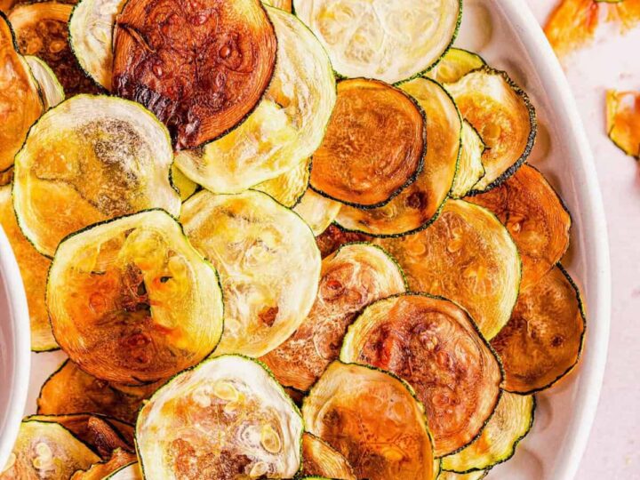 A white plate filled with crispy, golden-brown zucchini chips, some slightly darker than others, laid out in an overlapping pattern. The background is a light, neutral color with a few extra chips scattered around.