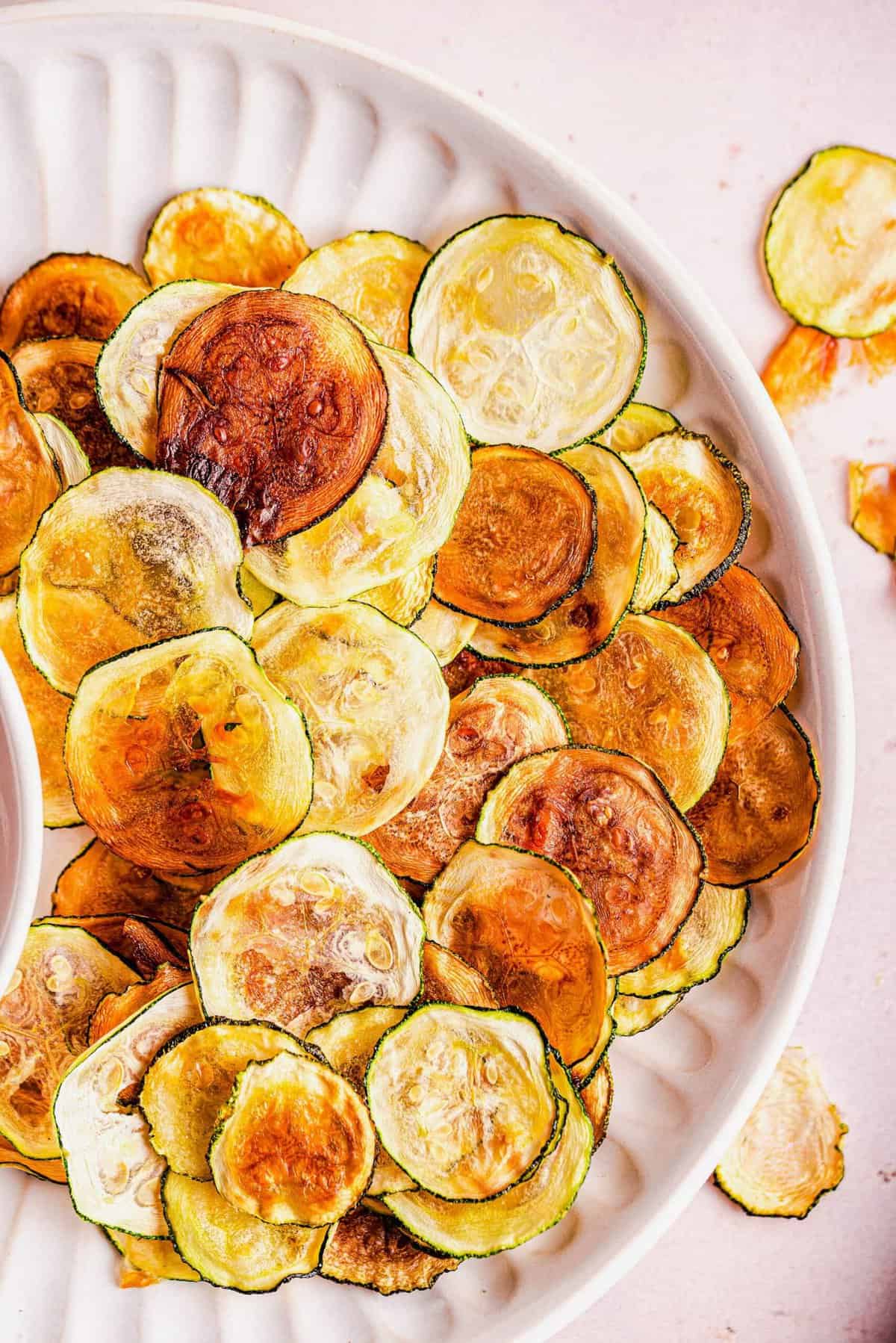 A white plate filled with crispy, golden-brown zucchini chips, some slightly darker than others, laid out in an overlapping pattern. The background is a light, neutral color with a few extra chips scattered around.