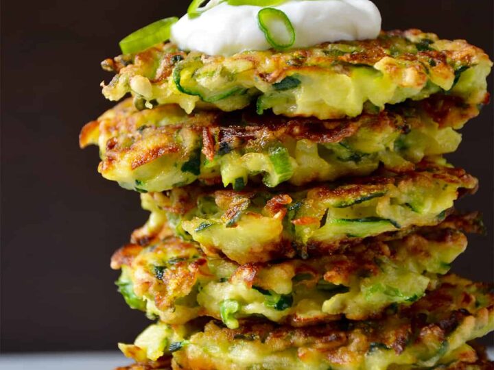 A stack of zucchini fritters is piled high on a white plate. The top fritter is garnished with a dollop of sour cream and thinly sliced green onions. The background is dark, highlighting the fritters golden-brown, crispy texture.
