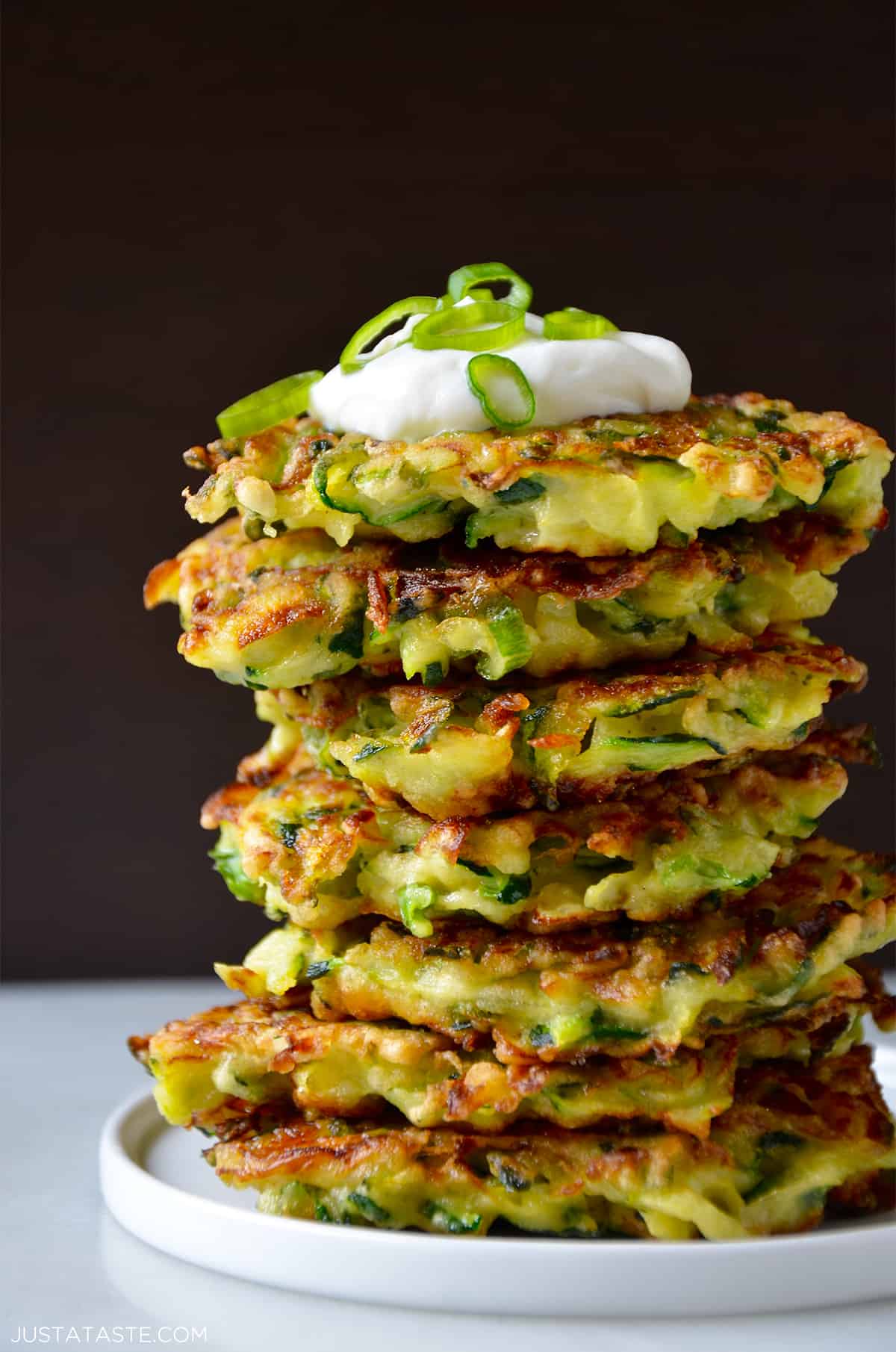 A stack of zucchini fritters is piled high on a white plate. The top fritter is garnished with a dollop of sour cream and thinly sliced green onions. The background is dark, highlighting the fritters golden-brown, crispy texture.