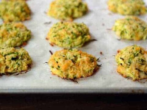Close-up of golden-brown baked zucchini fritters on a parchment-lined baking sheet. The fritters are evenly spaced and have a slightly crispy exterior, with visible shreds of zucchini throughout.