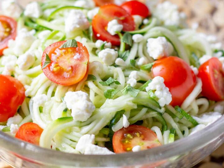 A glass bowl filled with zucchini noodles topped with halved cherry tomatoes, crumbled feta cheese, and chopped fresh herbs. The bowl is placed on a wooden surface.