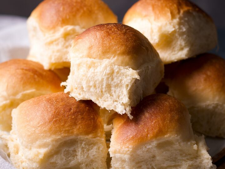 A plate of fluffy, golden-brown dinner rolls stacked on top of each other, showcasing their soft and textured interior under warm lighting.
