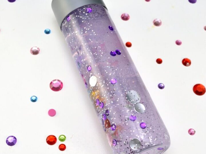A transparent bottle with a silver cap contains purple liquid and floating sequins, glitter, and shiny round embellishments. Colorful sequins are scattered on the white surface around the bottle.