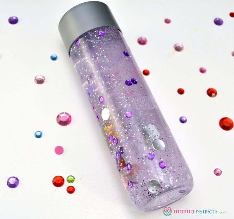 A transparent bottle with a silver cap contains purple liquid and floating sequins, glitter, and shiny round embellishments. Colorful sequins are scattered on the white surface around the bottle.