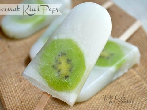 Close-up of a coconut popsicle with a slice of kiwi embedded within it, lying on a burlap surface. Other similar popsicles are visible in the background. The text reads Coconut Kiwi Pops and Will Cook for Smiles.