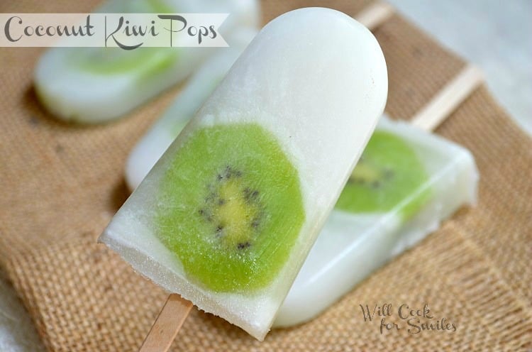 Close-up of a coconut popsicle with a slice of kiwi embedded within it, lying on a burlap surface. Other similar popsicles are visible in the background. The text reads Coconut Kiwi Pops and Will Cook for Smiles.