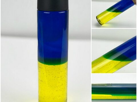 Tall clear bottle with black cap containing blue liquid on top and yellow liquid beneath, shown in various angles. The liquids remain separate, creating a distinct two-toned appearance.