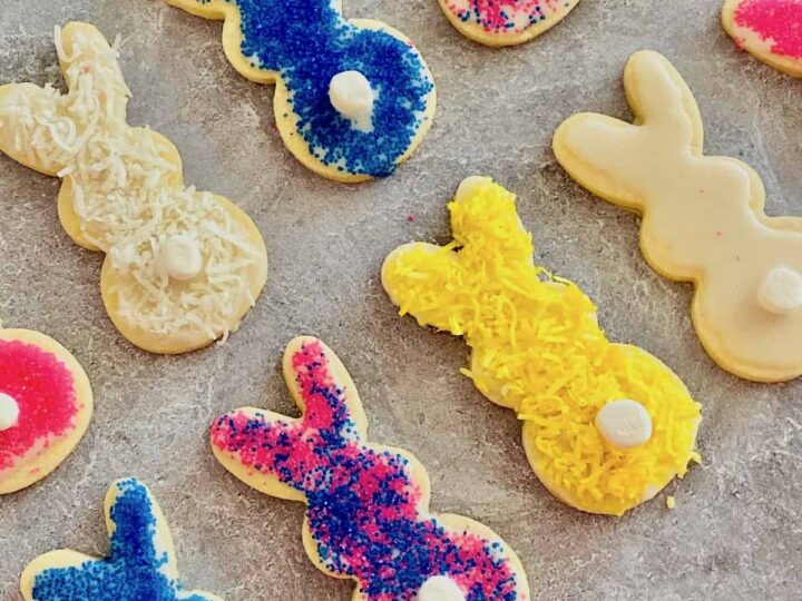 A variety of bunny-shaped cookies are decorated with colorful sprinkles and toppings. The cookies feature blue, pink, and yellow decorations with small white marshmallow tails on a gray surface.