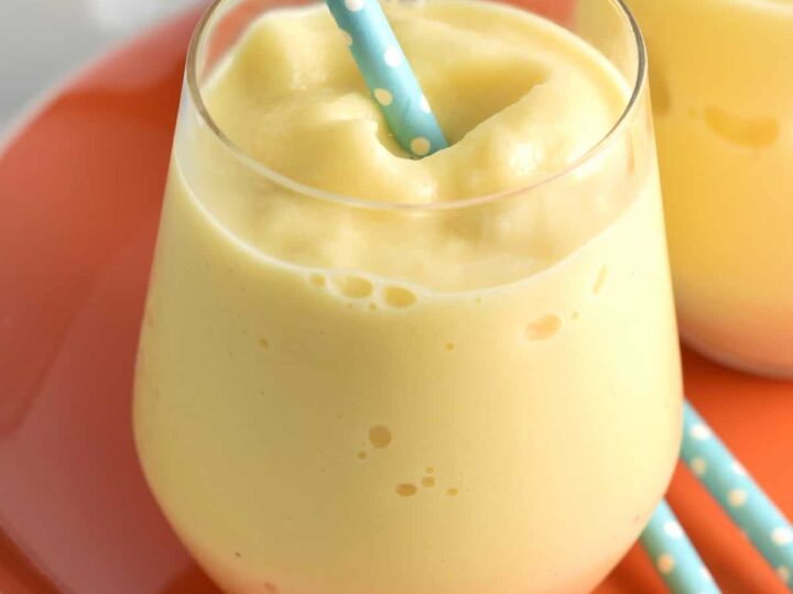 A creamy mango smoothie in a short glass with a blue polka dot straw on an orange plate. Another similar glass is partially visible in the background.