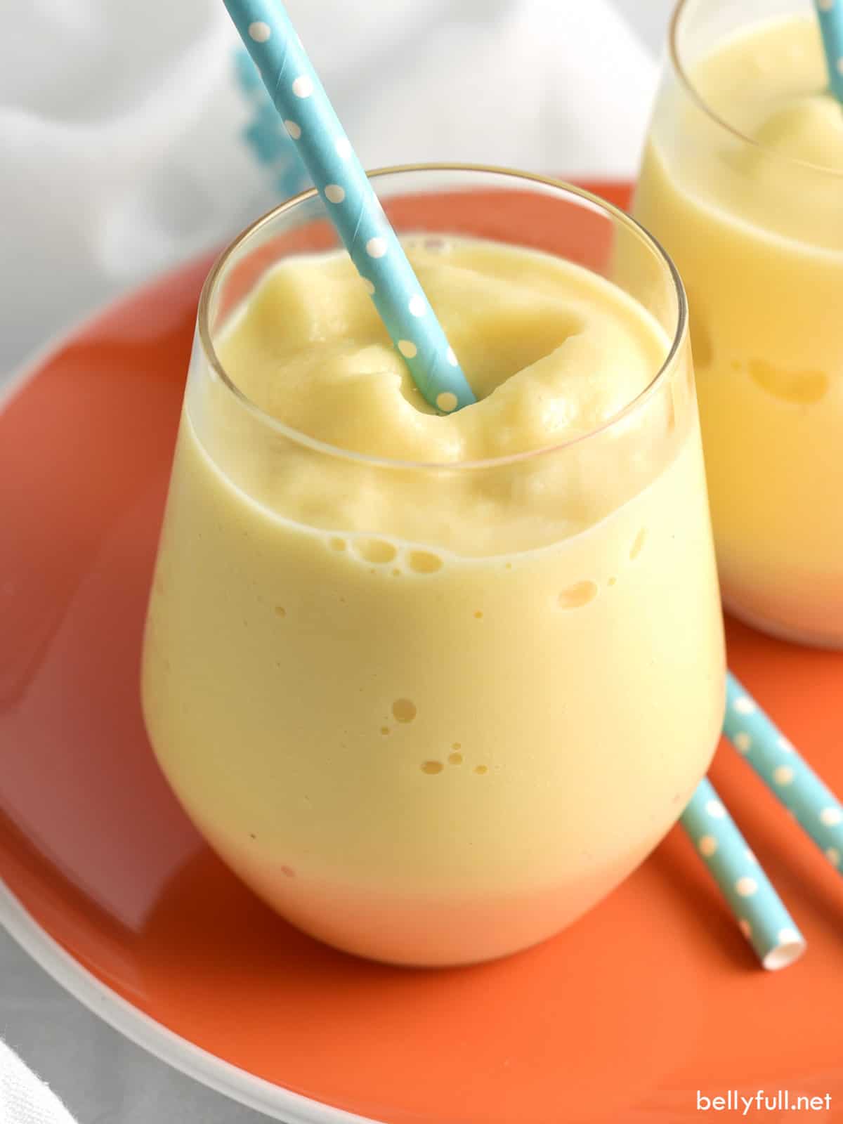 A creamy mango smoothie in a short glass with a blue polka dot straw on an orange plate. Another similar glass is partially visible in the background.